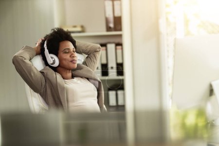 Téléchargez les photos : Femme d'affaires afro-américaine se détendre et écouter de la musique sur les écouteurs tout en profitant d'une pause pour étirer les mains derrière la tête dans un bureau seul au travail. - en image libre de droit