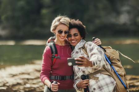 Foto de Dos amigas de raza mixta tomando selfies junto a un río durante una caminata por la montaña. - Imagen libre de derechos