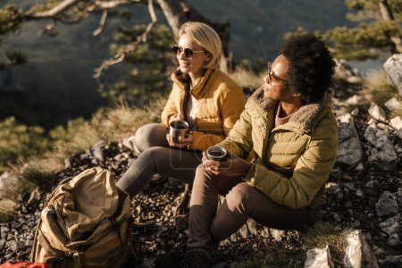 Foto de Dos amigas en la naturaleza disfrutando de una pausa para tomar café durante una caminata por las montañas. - Imagen libre de derechos