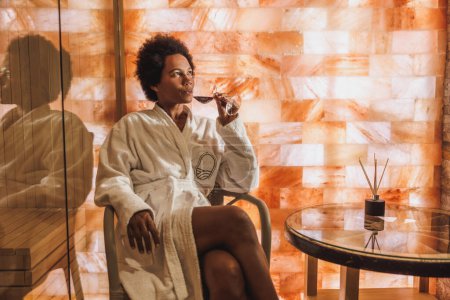 Photo for Beautiful mature African American woman drinking red wine and relaxing in a salt crystal cave sauna at the spa centre. - Royalty Free Image