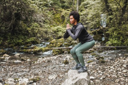 Téléchargez les photos : Belle coureuse afro-américaine mature faisant des exercices de squat après le jogging près de la rivière dans la nature. - en image libre de droit