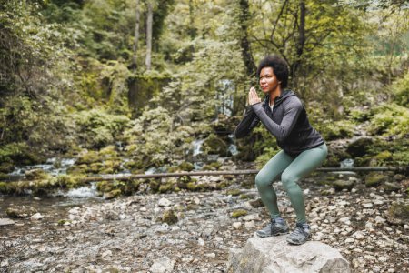 Photo for Beautiful mature African American female runner doing squat exercises near the river in nature. - Royalty Free Image