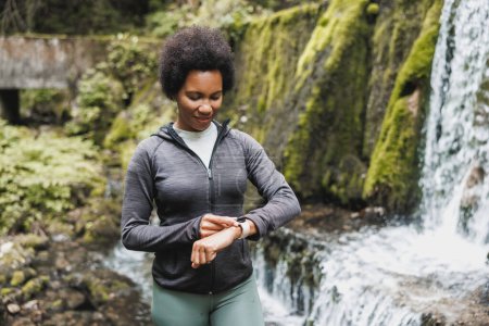 Téléchargez les photos : Femme mûre afro-américaine regarde smartwatch et se prépare à faire du jogging près de la cascade dans les montagnes. - en image libre de droit