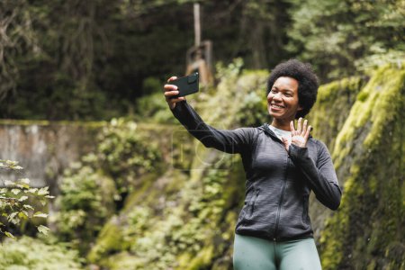 Photo for Mature African American woman taking selfie while standing near the waterfall and enjoying the view during her hike in the mountains. - Royalty Free Image