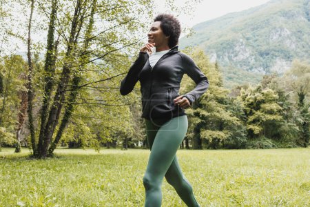 Téléchargez les photos : Belle souriante femme afro-américaine mature fait du jogging à travers l'herbe verte près du paysage de montagne. - en image libre de droit