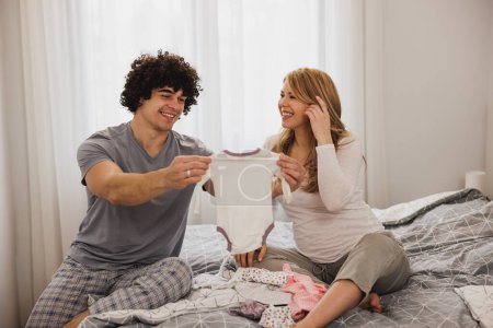 Photo for Happy pregnant couple looking at baby clothes while relaxing on a bed in bedroom. - Royalty Free Image