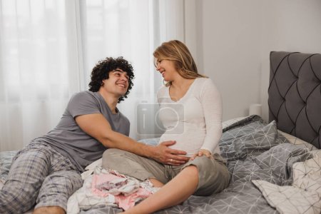 Téléchargez les photos : Joyeux couple enceinte parlant tout en se relaxant sur un lit dans la chambre. - en image libre de droit
