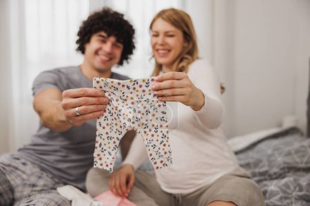 Téléchargez les photos : Joyeux couple enceinte regardant des vêtements de bébé tout en se relaxant sur un lit dans la chambre. - en image libre de droit