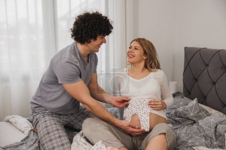 Téléchargez les photos : Joyeux couple enceinte tenant un vêtement de bébé tout en se relaxant sur un lit dans la chambre. - en image libre de droit