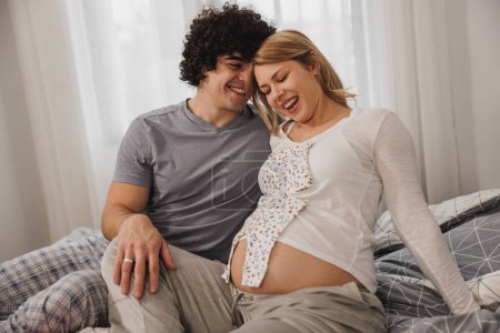 Téléchargez les photos : Heureux couple enceinte s'amuser avec des vêtements de bébé tout en se relaxant sur un lit dans la chambre. - en image libre de droit