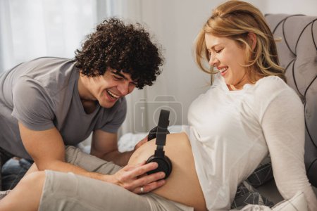 Téléchargez les photos : Couple enceinte s'amuser tout en se relaxant sur un lit dans la chambre. Père heureux tenant des écouteurs sur le ventre de sa femme enceinte. - en image libre de droit