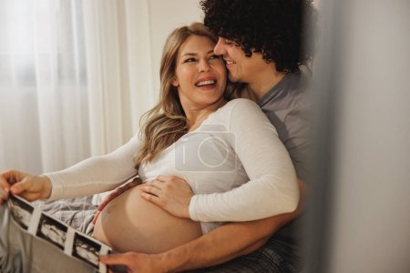 Téléchargez les photos : Heureux couple en attente de regarder le balayage médical de leur bébé tout en se relaxant sur un lit dans la chambre. - en image libre de droit