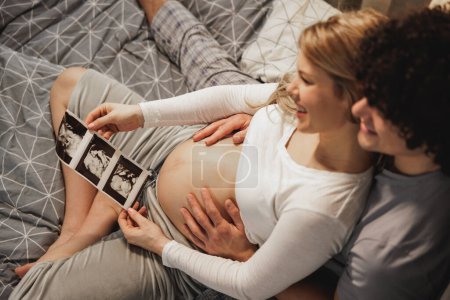 Foto de Feliz pareja embarazada mirando el escaneo médico de su bebé mientras se relaja en una cama en el dormitorio. - Imagen libre de derechos