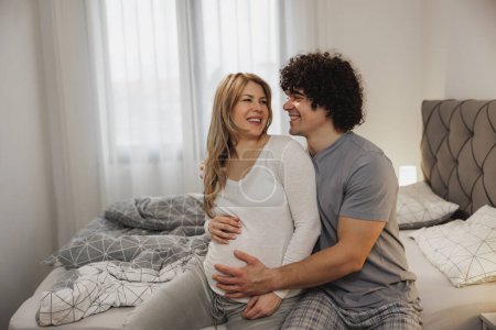 Téléchargez les photos : Heureux couple enceinte tenant la main sur un ventre enceinte tout en se relaxant sur un lit dans la chambre. - en image libre de droit