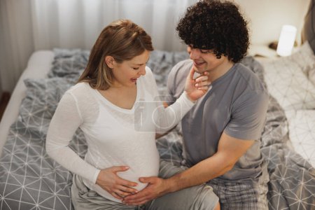 Téléchargez les photos : Heureux couple enceinte tenant la main sur un ventre enceinte tout en se relaxant sur un lit dans la chambre. - en image libre de droit
