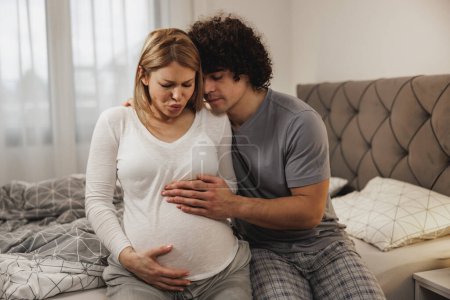 Téléchargez les photos : Homme inquiet consolant sa femme enceinte dans une chambre à coucher qui a des contractions prénatales. - en image libre de droit