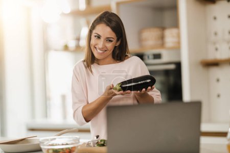 Photo for Shot of a young smiling woman preparing a healthy meal and making video blog on a laptop at home. - Royalty Free Image
