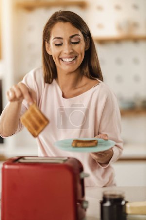 Téléchargez les photos : Tournage d'une femme souriante préparant le petit déjeuner pour elle-même, pain grillé dans la cuisine à la maison. - en image libre de droit