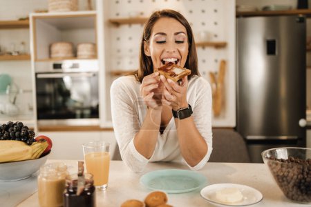 Téléchargez les photos : Tourné d'une jeune femme souriante prenant son petit déjeuner et profitant d'une matinée ensoleillée dans sa cuisine. - en image libre de droit