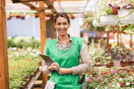 Foto de Hermosa mujer emprendedora sonriente está usando un delantal verde en un invernadero. Ella sostiene la tableta digital y mira a la cámara. - Imagen libre de derechos