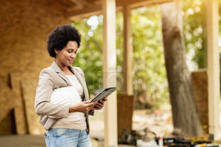 Photo for Shot of an African American female architect using a digital tablet and checking construction site of a new wooden house. - Royalty Free Image