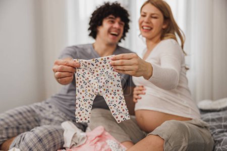 Téléchargez les photos : Joyeux couple enceinte montrant des vêtements de bébé tout en se relaxant sur un lit dans la chambre à coucher à la maison. - en image libre de droit