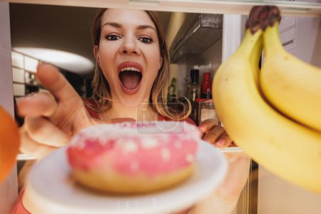 Foto de Sorprendido joven mujer mirando donas en el refrigerador durante la dieta. Enfoque selectivo. - Imagen libre de derechos