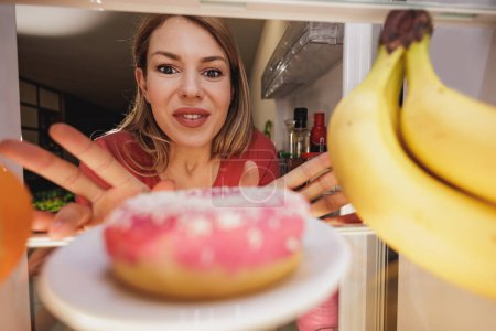 Foto de Mujer joven emocionada mirando donas en el refrigerador durante la dieta. Enfoque selectivo. - Imagen libre de derechos