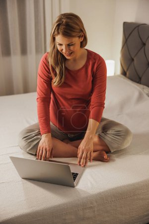 Foto de Relajado joven futura madre haciendo compras en línea en el ordenador portátil mientras se relaja en una cama en el dormitorio. - Imagen libre de derechos