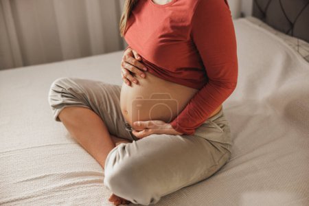 Téléchargez les photos : Gros plan d'une femme enceinte méconnaissable tenant son estomac tout en se relaxant sur un lit dans la chambre. - en image libre de droit