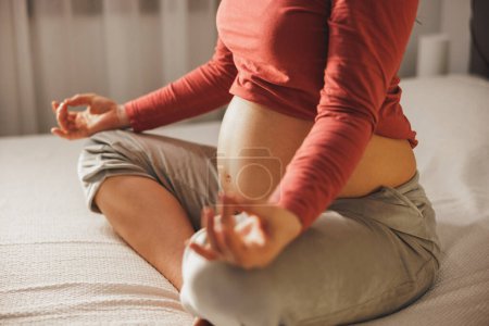 Foto de Primer plano de una mujer embarazada irreconocible haciendo ejercicios de meditación de yoga en posición de loto en una cama en el dormitorio. - Imagen libre de derechos