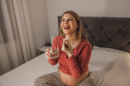 Téléchargez les photos : Belle future mère tenant un verre d'eau et prenant une pilule vitaminée sur un lit dans la chambre. - en image libre de droit
