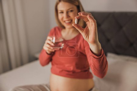 Téléchargez les photos : Gros plan d'une femme enceinte tenant un verre avec de l'eau et buvant des médicaments sur un lit dans la chambre. - en image libre de droit