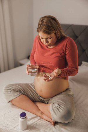 Téléchargez les photos : Froncement de sourcils jeune femme enceinte se sentant mal, obtenir de l'aspirine ou un analgésique sur un lit dans la chambre. - en image libre de droit