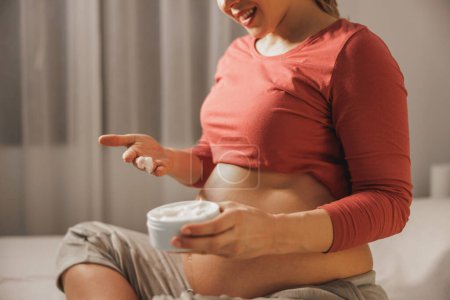 Foto de Primer plano de una mujer embarazada irreconocible aplicando crema hidratante en su estómago en el dormitorio. Evitar la marca de estiramiento. - Imagen libre de derechos