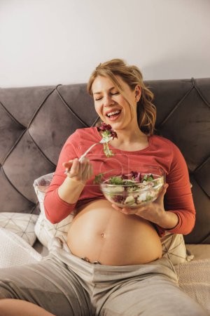 Foto de Feliz mujer embarazada comiendo ensalada de verduras frescas mientras está sentada en la cama en su dormitorio. - Imagen libre de derechos