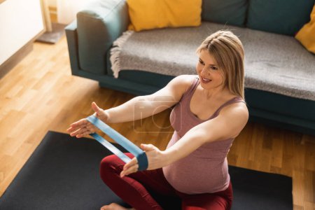 Téléchargez les photos : Jeune femme enceinte faisant de l'exercice avec élastique à la maison. Elle fait un entraînement d'étirement dans son salon. - en image libre de droit