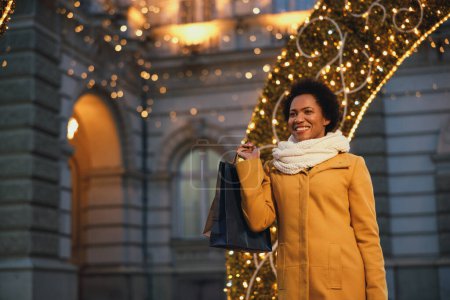Téléchargez les photos : Belle femme noire souriante marchant et faisant du shopping pendant les vacances de Noël dans la ville. - en image libre de droit
