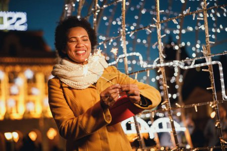 Photo for Middle age black woman holding sparklers and Christmas gift at festive winter night in the city. - Royalty Free Image