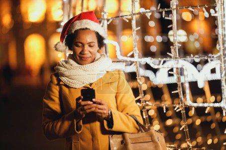 Foto de Mujer negra de mediana edad surfeando en las redes sociales en su smartphone en la festiva noche de Navidad en la ciudad. - Imagen libre de derechos