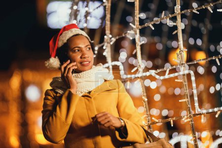 Foto de Mujer negra de mediana edad hablando en su teléfono inteligente en la noche de Navidad festiva en la ciudad. - Imagen libre de derechos