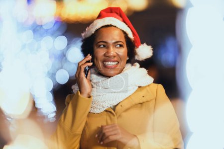 Foto de Mujer negra de mediana edad hablando por teléfono inteligente en el mercado de Navidad en la noche festiva en la ciudad. - Imagen libre de derechos