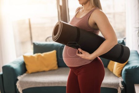 Foto de La toma de una mujer embarazada irreconocible sosteniendo una esterilla de ejercicio está lista para practicar yoga en casa por la mañana.. - Imagen libre de derechos