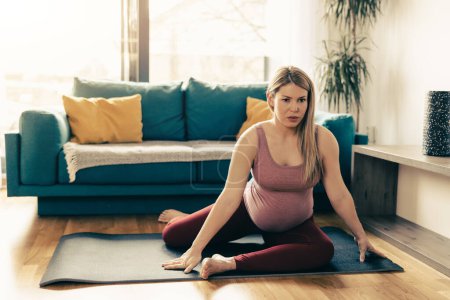Téléchargez les photos : Jeune femme enceinte exerçant la mobilité à la maison le matin. Elle s'entraîne sur un tapis d'exercice et s'étire dans son salon. - en image libre de droit