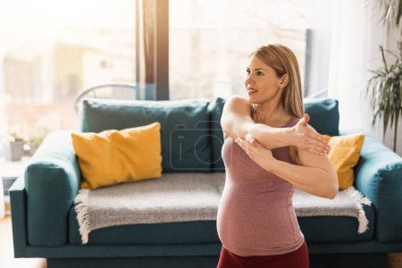 Photo for A pregnant woman in sports clothing is stretching and exercising to promote wellbeing in her living room. - Royalty Free Image