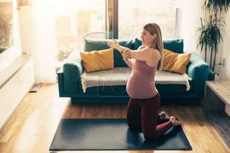 Téléchargez les photos : Jeune femme enceinte faisant de l'exercice à la maison. Elle s'entraîne sur un tapis d'exercice et s'étire dans son salon. - en image libre de droit