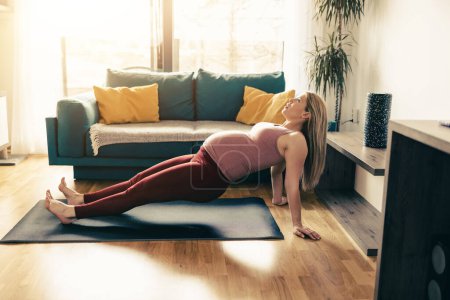 Foto de Mujer embarazada joven haciendo yoga en casa por la mañana. Ella está haciendo ejercicios de estiramiento. - Imagen libre de derechos