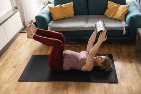 Téléchargez les photos : Jeune femme enceinte faisant de l'exercice à la maison le matin. Elle s'entraîne sur un tapis d'exercice et fait de l'exercice de mobilité dans son salon. - en image libre de droit