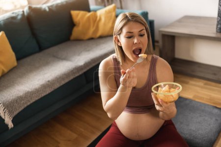 Foto de Feliz joven embarazada comiendo ensalada de frutas frescas mientras hace ejercicio en casa. Ella se relaja en la alfombra de ejercicio en su sala de estar por la mañana. - Imagen libre de derechos