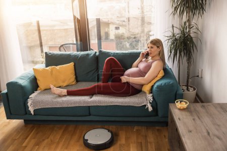 Téléchargez les photos : Jeune femme enceinte se détendre sur le canapé à la maison le matin tandis que l'aspirateur robot travaille et nettoie le salon. - en image libre de droit